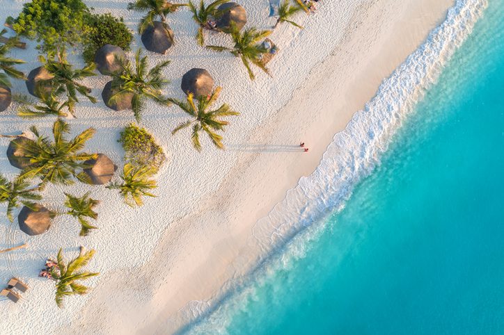Aerial view of umbrellas, palms on the sandy beach of Indian Ocean at sunset. Summer holiday in Zanzibar, Africa. Tropical landscape with palm trees, parasols, white sand, blue water, waves. Top view VolunteerForever